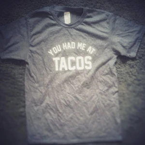 Grå t-shirt med text från Gildan - Grå t-shirt med vit tryckt text 'You had me at TACOS' på bröstet. Klassisk rund hals och korta ärmar. Perfekt för dig som älskar tacos och vill ha en chill och rolig stil.