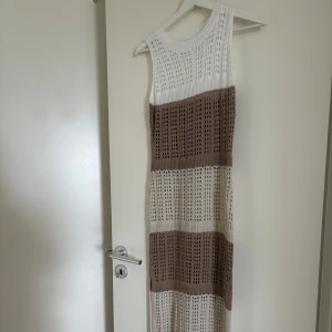 Stickad långklänning i vitt och beige - Säljer en stickad långklänning med breda ränder i vitt och beige. Klänningen är ärmlös och har en rund halsringning. Perfekt för varma dagar och enkel att styla med sandaler eller sneakers.