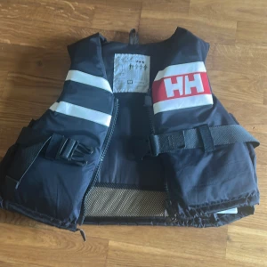 Marinblå flytväst från Helly Hansen - Säljer en marinblå flytväst från Helly Hansen med vita reflexdetaljer och röd HH-logga. Västen har justerbara spännen och bälte runt midjan. Perfekt för aktiviteter på vattnet eller båtliv.