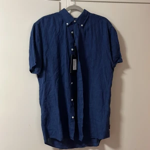 Blå linneskjorta från Bondelid - Snygg blå skjorta i linne från Bondelid med korta ärmar och klassisk krage. Skjortan har vita knappar framtill och en avslappnad passform, perfekt för varma dagar. Ny med prislapp. Nypris 599