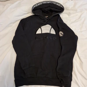 Sommar25. Svart hoodie från Ellesse med broderad logga - Säljer en svart hoodie från Ellesse med stor broderad logga på bröstet och patch på ärmen med texten 'Cortina D'Ampezzo'. Huva med dragsko och tryckt Ellesse-mönster på insidan. Perfekt för en sportig och avslappnad stil.