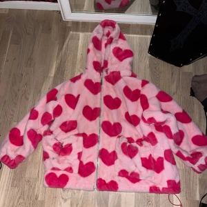 Rosa pälsjacka med hjärtan från Allured - Supermysig rosa pälsjacka med stora röda hjärtan och huva från Allured. Jackan har dragkedja framtill och två fickor. Perfekt statement-plagg för dig som vill sticka ut och hålla dig varm.