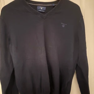 Svart v-ringad tröja från GANT - Klassisk svart v-ringad tröja från GANT i mjuk bomull. Diskret GANT-logga broderad på bröstet. Perfekt basplagg med ribbade muddar vid hals, ärmslut och nederkant.