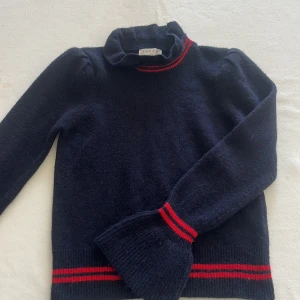 Mörkblå stickad tröja med röd rand från Sandro - Mörkblå stickad tröja från Sandro Paris med röd rand vid krage, ärmslut och nederkant. Tröjan har hög, volangig krage och långa ärmar. Perfekt för dig som gillar klassiska detaljer med en twist. Den är i sandro storleken 1 och är supersöt nu till sommaren med en vit kjol!! Aldrig använd 