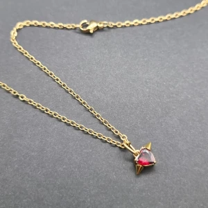 Guldhalsband med röd hjärtberlock - Snyggt halsband i guldfärgad metall med en kedja och ett hängsmycke format som ett hjärta i röd sten. Berlocken har två små guldfärgade spikes på sidorna som ger en cool edge. Perfekt för dig som gillar unika detaljer och färg.