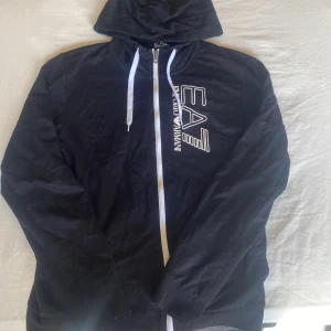 Svart hoodie från Emporio Armani - Svart hoodie från Emporio Armani med vit dragkedja och vita snören i huvan. Stort EA7-tryck i vitt på bröstet ger en cool look. Tröjan har långa ärmar och är tillverkad i mjuk bomull, perfekt för chill dagar. Snygg och enkel att matcha med jeans eller joggers.