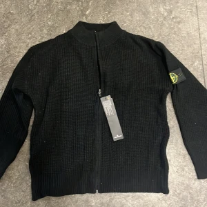 Svart stickad kofta från Stone Island - Svart stickad kofta från Stone Island med dragkedja framtill och klassisk logga på ärmen. Tröjan har ribbad struktur, hög krage och långa ärmar. Perfekt för dig som gillar stilrena och tidlösa plagg med streetkänsla. Den är storlek L men passar även M. Nytt skick!