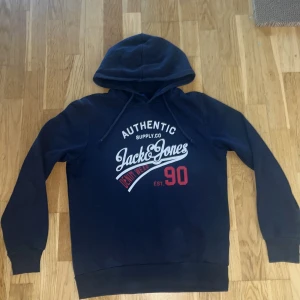 Mörkblå hoodie Jack & Jones storlek S - Snygg mörkblå hoodie från Jack & Jones med vit och röd print på bröstet. Tröjan har huva med snörning och långa ärmar. Perfekt för chill dagar och streetstyle. Mjuk insida och klassisk passform. Ikonisk logga framtill för extra stilpoäng. Tröjan är i storlek S och är i mycket bra skick. Har du några funderingar kom skriv dm.