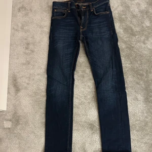 Mörkblå jeans från Nudie Jeans - Snygga mörkblå skinny jeans från Nudie Jeans med klassiska orange sömmar och femficksdesign. Jeansen har en smal passform och är tillverkade i stretchig denim för extra komfort. Perfekta för dig som gillar en stilren och modern look. Storlek W27 L30