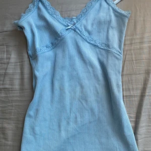 Ljusblå ribbad linnetopp H&M Divided - Supersöt ljusblå ribbad linnetopp från H&M Divided med spetskant och liten rosett framtill. Tunt, stretchigt material och smala axelband. Perfekt för lager-på-lager eller till jeans. Sitter tight och har en feminin v-ringning med spetsdetaljer. Aldrig använt den 💕