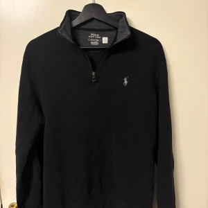 Svart half zip tröja från Polo Ralph Lauren - Svart långärmad tröja från Polo Ralph Lauren med half zip och klassisk krage. Tröjan har en broderad logga på bröstet och är gjord i mjuk bomull. Perfekt för chill dagar eller när du vill ha en clean look. Ganska kort i magen och lite pösig tröja så om du gillar när tröjan är slimmad mot kroppen eller inte är lite biffig är denna tröja inget för dig
