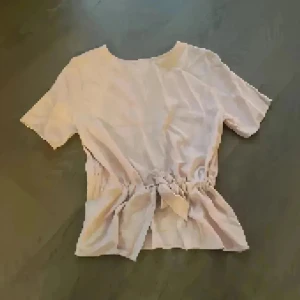 Ljusrosa blus från Selected Femme - Supersöt ljusrosa blus från Selected Femme med korta ärmar och rund hals. Midjan har snygg knytning som ger en feminin siluett. Blusen är i ett mjukt, lätt material och passar perfekt till både jeans och kjol.