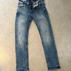 Slim fit jeans från Jack & Jones - Ljusblå slim fit jeans från Jack & Jones, modell Glenn. Jeansen har slitna detaljer, klassisk femficksdesign och knäppning med knappar. Snygg tvätt med lätt urtvättade partier och bekväm passform som sitter tajt men ändå flexibelt.