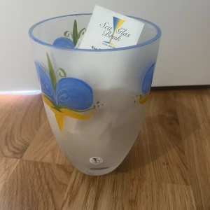 Oanvänd Sea Glas Bruk - Handmålad vas i frostat glas från Sea Glas Bruk, designad av Lena Engman. Vasen har ett färgglatt blommotiv i blått och gult, Svenskt hantverk från Småland. Helt oanvänd och har legat i originalförpackning hela tiden. Kartongen är lite sliten efter förvaring men innehållet är i perfekt skick. 📦 Kan skickas på köparens ansvar (packas noggrant) eller hämtas i handen. 📩 Skicka gärna ett meddelande vid frågor eller om du vill ha flera bilder! 🌸😊 📦 Jag skickar snabbt och packar alltid med omsorg!