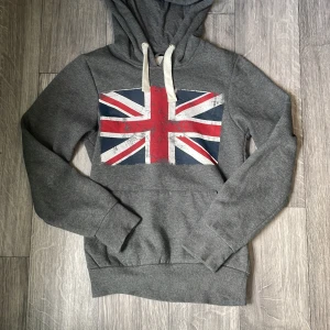 Grå hoodie med Union Jack från Cedar Wood State - Snygg grå hoodie från Cedar Wood State i storlek XS. Stor brittisk flagga (Union Jack) framtill, vita snören i huvan och klassisk känguruficka. Perfekt för dig som gillar streetstyle och vill sticka ut lite extra.