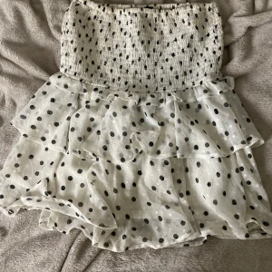 Prickig volangklänning från Lindex XS - Supersöt kort klänning från Lindex i storlek XS. Klänningen är vit med svarta prickar, har smock upptill och flera lager volanger nedtill. Axelbandslös design och luftig känsla, perfekt för varma dagar. Materialet är lätt och känns som bomull.