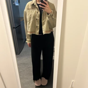  croppad jeansjacka från Zara - Säljer en croppad beige jeansjacka från Zara med broderade detaljer i rosa, svart och grönt. Jackan har rå kant nertill, knappar framtill och på ärmslut samt snygga broderier längs krage, ärmar och nederkant. Perfekt för dig som vill sticka ut lite extra.