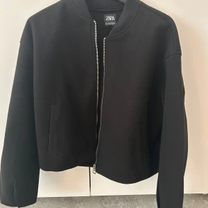 Svart bomberjacka från Zara - Säljer en svart bomberjacka från Zara med stilren design och rak passform. Jackan har ribbad krage, långa ärmar och en tydlig dragkedja framtill. Perfekt för dig som gillar en clean och enkel look.