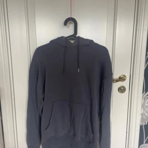 Mörkgrå hoodie från Jack & Jones - En stilren mörkgrå hoodie från Jack & Jones i storlek S. Tröjan har huva med snörning och en stor magficka framtill. Perfekt för dig som gillar en enkel och avslappnad look. Tillverkad i mjuk bomullsmix för extra komfort.