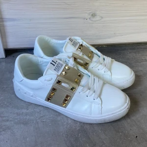 Valentino Garavani Rockstud sneakers - HELT NY snygga vita/guld sneakers från Valentino Garavani med ikoniska guldiga nitar på ett beige band över vristen. Skorna har rund tå, vita snören och är tillverkade i skinn. Perfekta för dig som vill sticka ut med exklusiva detaljer och lyxig känsla.