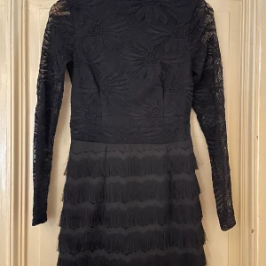 Svart långärmad spetsklänning med fransar - Supersnygg svart klänning med långärmade spetsdetaljer och blommigt mönster upptill. Kjolen har flera lager med fransar som ger cool rörelse. Perfekt för dig som vill sticka ut med en unik look och älskar detaljer!