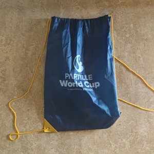 Blå Partille World Cup ryggsäck - Säljer en blå gympapåse/ryggsäck från Partille World Cup med gula snören och vit logga. Perfekt för att bära träningskläder eller skor. Enkel och smidig design med snörning upptill och förstärkta hörn.
