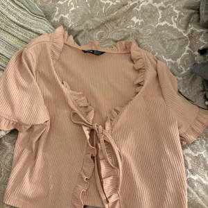 Ljusrosa ribbad blus med volang - Supersöt ljusrosa blus från SHEIN med ribbad struktur och volang längs v-ringningen. Knyt framtill ger en extra gullig touch. Perfekt för dig som gillar romantisk stil och vill ha något som sticker ut lite extra.