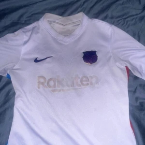 Vit FC Barcelona Nike fotbollströja strö 147/158 - Snygg vit FC Barcelona fotbollströja från Nike med klubbmärke och Nike-logga på bröstet. Rakuten och Unicef-loggor tryckta på fram- och baksidan. Tröjan är kortärmad och tillverkad i Dri-FIT-material som andas. Perfekt för dig som älskar Barca och vill ha en clean look.