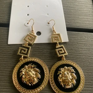 Statement örhängen med lejonmotiv - Säljer ett par riktigt coola statement örhängen i guld och svart. De har två fyrkantiga spiralformade detaljer och stora runda hängen med ett lejonhuvud i guld på svart bakgrund. Perfekta för dig som vill sticka ut och addera lite edge till din look.