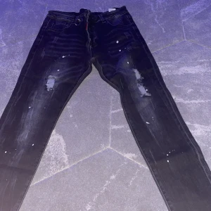 Svarta slim jeans från Dsquared2 - Säljer ett par svarta slim fit jeans från Dsquared2 med coola slitningar och vita färgstänk. Jeansen har klassisk femficksdesign och är tillverkade i Italien. Materialet är jeans och passformen är slim, perfekt för dig som gillar en modern look.