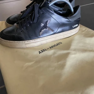 Svarta sneakers från Axel Arigato - Snygga svarta sneakers från Axel Arigato med broderad fågel på sidan och kontrasterande beige sula. Skorna är handgjorda i Portugal och är tillverkade i läder med detaljer i mocka. En dustbag tillkommer med skorna. Nypris: 2750kr. Mitt pris: 950kr. Använda med i välbehållet skick. Har några få slitningar (se bild 5) därav priset. Har du frågor eller funderingar, tveka inte på att höra av dig!