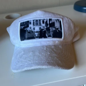 Vit velvetkeps  - Snygg vit emilio pendenza  velvetkeps med tryck av ett par människor samt byggnader.  Den är i väldigt bra skick förutom att det är lite beige märken i kepsen men som jag ska tvätta bort så gott det går!