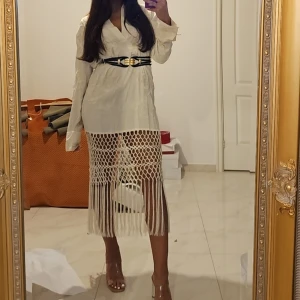 Vit långklänning med fransar och bälte - Säljer en vit långklänning med coola fransdetaljer och nätmönster nedtill. Klänningen har lång ärm, v-ringning och ett svart bälte med gulddetaljer i midjan. Perfekt för dig som vill sticka ut med en unik look.