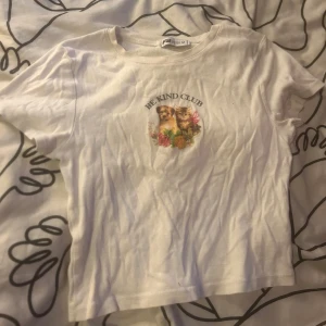 Vit t-shirt med tryck - Vit t-shirt från Sister med korta ärmar och ett gulligt tryck framtill med texten 'Be Kind Club', katt, hund och blommor. Klassisk passform och rund halsringning. Perfekt för dig som gillar söta motiv och vill ha en chill vibe.