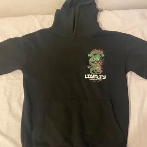 Svart hoodie med draktryck Paranoid - Svart hoodie från Paranoid med stor, färgglad drake tryckt på ryggen och mindre drakmotiv framtill. Texten 'LOYALTY' och 'PARANOID' i vitt ger en edgy vibe. Hoodien har huva och magficka, perfekt för dig som gillar streetwear med asiatiska influenser.