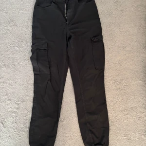Svarta cargopants med fickor - Svarta cargopants med stora sidofickor och dragkedja framtill. Byxorna har en avslappnad passform och resår vid bensluten.☺️