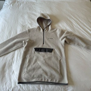 Beige hoodie från Columbia - Säljer en beige hoodie från Columbia med huva och half zip. Tröjan har en stor magficka och liten ficka ovanför där du kan lägga plånbok eller nycklar för säkrare förvaring. 