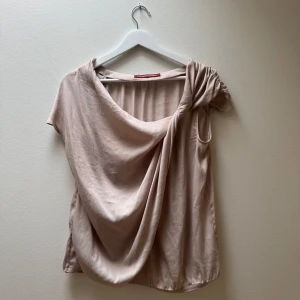 Omlottopp Comptoir des Cotonniers - Säljer en superfin beige/rosa omlottopp från Comptoir des Cotonniers. Toppen har kort ärm, draperad front och snygg knutdetalj på axeln. Mjuk och lyxig känsla, perfekt för dig som gillar en unik och elegant look.