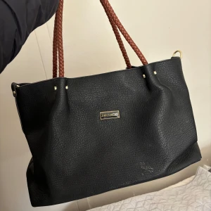 Svart axelväska - Stilren svart axelväska från Jimmy C med bruna flätade axelband och guldfärgade detaljer. Väskan är rymlig med flera innerfack och dragkedja. Tillverkad i skinnimitation med en klassisk, enkel design som passar till det mesta. (ej äkta)