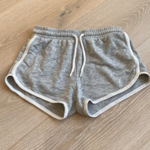 Grå sportiga shorts med snörning - Grå sportshorts med vit kant och snörning i midjan. Mjukt bomullsmaterial och elastisk midja för skön passform. Perfekta för sommarens alla aktiviteter eller bara att chilla i. Klassisk sportig look med rundade benkanter.