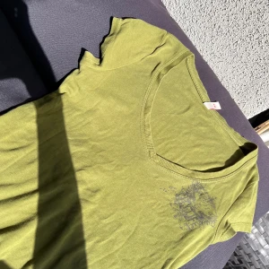 Grön v-ringad t-shirt S.Oliver XS - Snygg grön t-shirt från S.Oliver i storlek XS. T-shirten har v-ringning, korta ärmar och ett tryck med text och emblem på bröstet. Tillverkad i mjuk bomull, perfekt för en chill och avslappnad stil.Cool tshirt i bra kvalitet med snygg detalj och v-ringning. Väldigt bra material. Fin färg, mörkare grön nästan lite militär färg.  