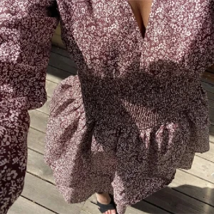 Vinröd/lila blommig kortklänning med v-ringning - Supersöt vinröd/lila kortklänning med vitt blommigt mönster, v-ringad hals och långa puffärmar. Klänningen har smock i midjan som ger en snygg siluett och voluminös kjol. Perfekt för dig som gillar romantiska detaljer och vill sticka ut.