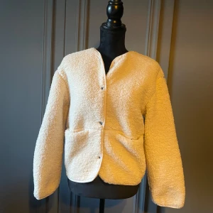 Beige teddyjacka från H&M - Supermysig beige teddyjacka från H&M med stora fickor framtill och tryckknappar. Jackan har en rak, loose passform och är tillverkad i fluffigt syntetmaterial som ger en cool och avslappnad vibe. Är i XXS men passar mig som bär M.