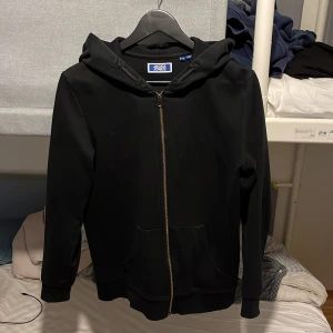Svart hoodie från Jack & Jones - Svart hoodie från Jack & Jones med dragkedja framtill och två fickor. Klassisk modell med huva och långa ärmar, perfekt för chill dagar. Tillverkad i mjukt material som känns skönt mot huden.