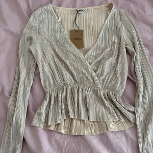 Beige omlottopp med volang - Snygg champagnefärgad omlottopp med plisserat tyg och glansig finish. Toppen har långa ärmar, v-ringning och volang nedtill som ger en feminin touch. Perfekt för dig som vill sticka ut med en elegant och trendig look.