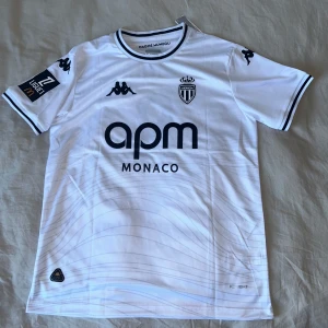 AS monaco 24-25 - AS Monaco fotbollströja i vitt med svarta detaljer, Pogba och nummer 8 på ryggen. Kappa-logga på axlarna, klubbmärke på bröstet och Ligue 1-märke på ärmen. Tillverkad i lätt och ventilerande polyester, perfekt för match eller träning.