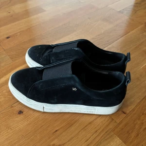 Svarta mockasneakers från KCobler - Stilrena svarta sneakers från KCobler i mjuk mocka med bred resår över vristen för enkel påtagning. Skorna har vit platt sula och diskret logga på sidan. Perfekta för en clean och avslappnad look.