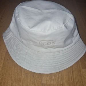 Vändbar bucket hat från Egon - Snygg vändbar bucket hat från Egon. Ena sidan är helvit med diskret logga, andra sidan har ett coolt mönster med stora bokstäver i ljusgrönt och vitt. Hatten är i mjuk bomull och har klassisk form med bred brätte. Perfekt för dig som vill variera din stil.