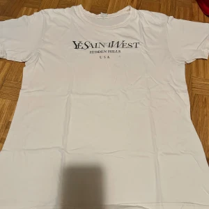 Vit YeSaintWest t-shirt XL - Vit t-shirt från YeSaintWest i storlek XL med tryck 'Hidden Hills USA' på bröstet. Klassisk rund hals och korta ärmar. Tillverkad i mjuk bomull, perfekt för chill dagar. Snygg och enkel design som funkar till jeans eller shorts.