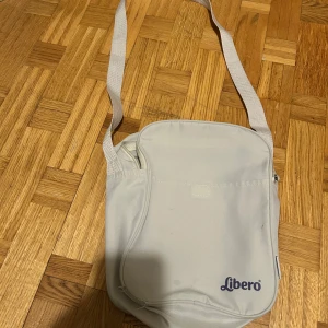 Vit axelväska från Libero - Säljer en stilren vit axelväska från Libero med justerbar rem och dragkedja upptill. Väskan har en enkel design med ett stort huvudfack och är tillverkad i slitstark polyester. Perfekt för att bära med sig det viktigaste till skolan eller stan.